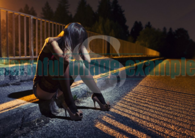 A woman in a tank top and shorts, with knee high stockings and heels. She's sitting on the side of the road at night, with there head in her hands and hair covering her face. White SWAN logo with text reads: example harmful image.