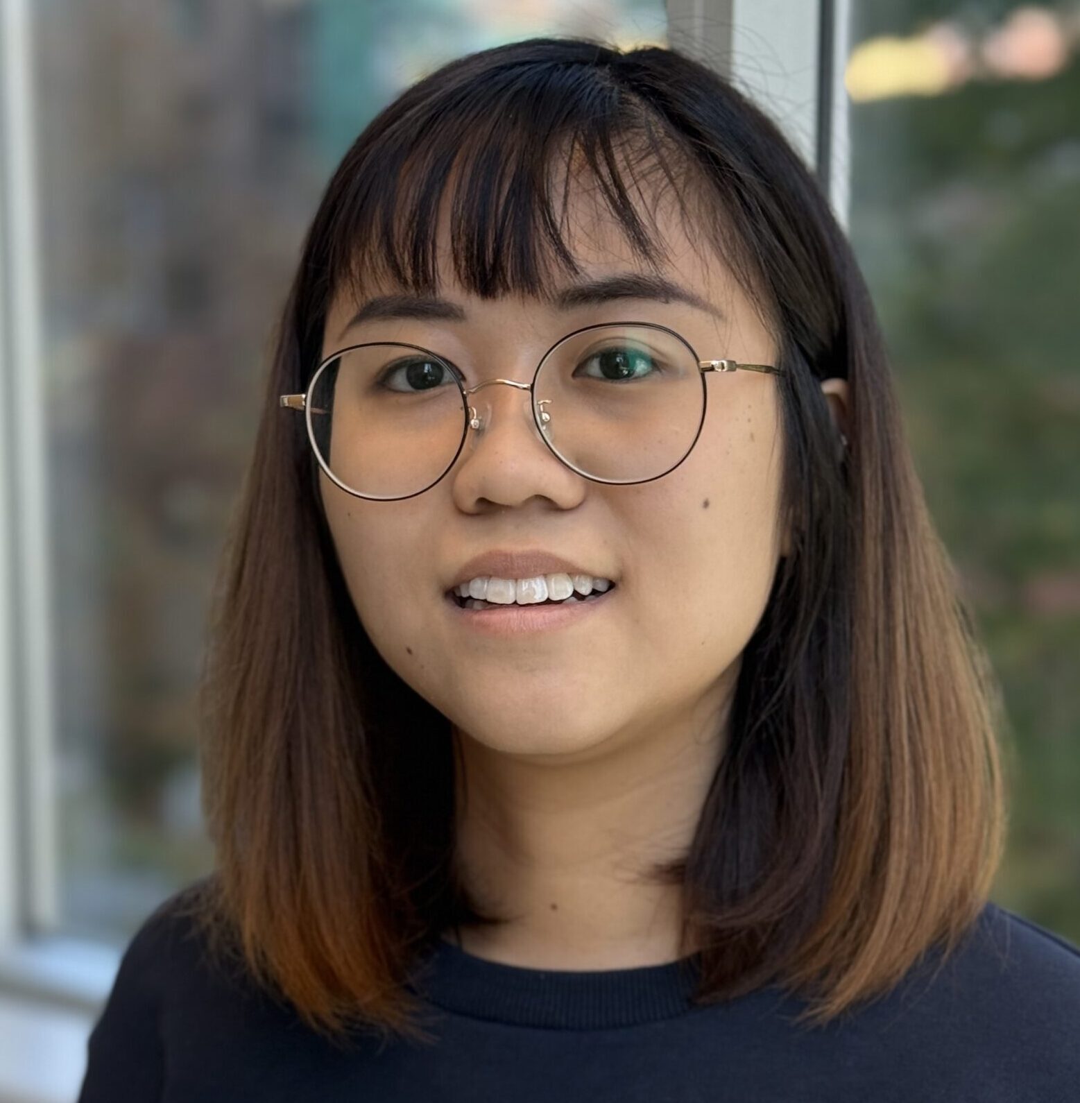 Chantel in the centre smiling, wearing glasses and a checkered shirt. Window showing nearby apartments in the background.